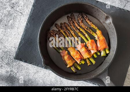 Spargel in Rückenspeck eingewickelt, in einer gusseisernen Bratpfanne gekocht. Auf einem Schiefer Schneidebrett Stockfoto