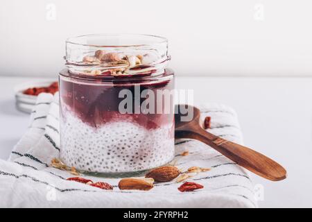 Reich an Mikronährstoffen Chia Pudding in Glas mit Kokosmilch, Marmelade, Müsli, Samen und Nüsse. Cremig roh veganes Frühstücksdessert auf gestreifter Serviette Stockfoto