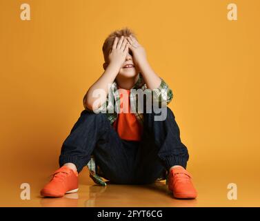 Blonde lächelnde junge Kind in karierten Hemd und Turnschuhe sitzen Und die Augen mit Händen bedecken Stockfoto