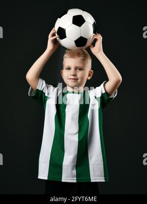 Blonde lächelnde Jungen Kind in Sport grün und weiß Kleidung Im Stehen den Fußball über den Kopf halten Stockfoto