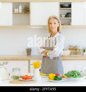 Porträt einer schönen, positiv reifen Frau in der Schürze in der Küche, die die Kamera ansah und lächelte, einer pensionierten Großmutter, die neben dem Tisch mit gesunden Zutaten stand, während sie für die Familie zu Hause kochte Stockfoto