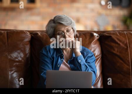 Durchdachte, reife Frau, die mit einem Laptop auf dem Sofa sitzt Stockfoto