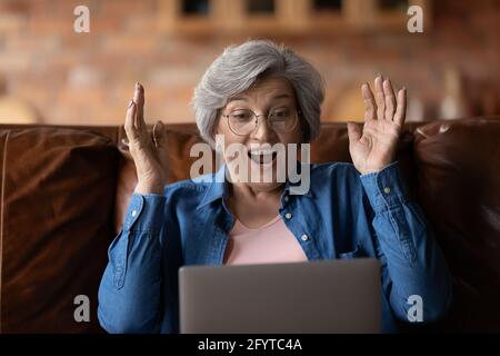 Reife Frau mit offenem Mund lesen unglaubliche Nachrichten auf Laptop Stockfoto