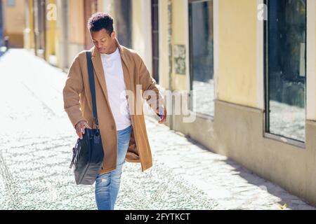 Ein junger schwarzer Mann, der die Straße entlang läuft, trägt eine Aktentasche und ein Smartphone. Stockfoto