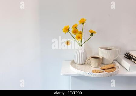 Ein Bouquet von Delelionen in einer weißen, geriffelten Vase und zwei Tassen in verschiedenen Größen mit Kaffee auf einem weißen Tisch. Frühstück. Speicherplatz kopieren Stockfoto