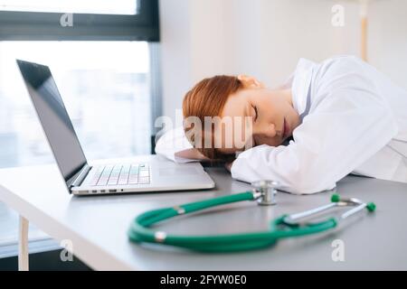 Müde erschöpfte, überarbeitete junge Ärztin im weißen Mantel, die am Schreibtisch mit Laptop und Stethoskop am Arbeitsplatz schlief. Stockfoto