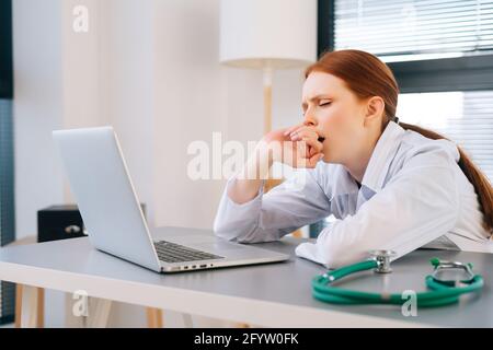 Nahaufnahme der müden, erschöpften jungen Ärztin mit weißem Mantel, der während der Arbeit am Laptop gähnt. Stockfoto
