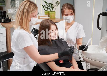 Horizontale Momentaufnahme einer Zahnärztin und Assistentin, die dem Patienten in der Klinik den Röntgenbericht erklärt. Junges Mädchen im Zahnarztstuhl, trägt Lätzchen, hört dem Arzt zu. Zahnmedizin Konzept Stockfoto