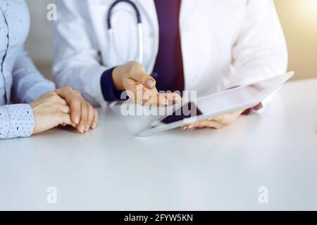 Unbekannte Ärztin zeigt ihrem Patienten eine Beschreibung der Medikamente, während sie zusammen am Schreibtisch in der sonnigen Vitrine in einer Klinik sitzt Stockfoto