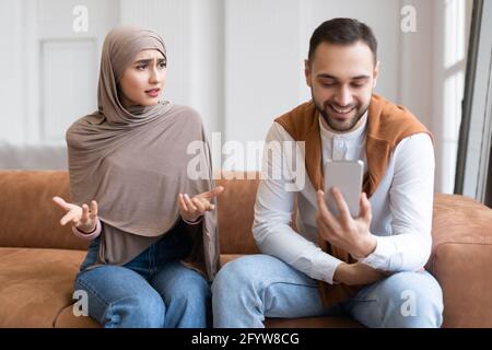 Unzufrieden Muslimische Frau Beim Blick Auf Ehemann Texting Auf Smartphone Indoor Stockfoto