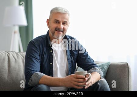 Schöner grauhaariger älterer Mann, der im Wohnzimmer Kaffee trinkt Stockfoto