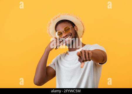 Fröhlicher schwarzer Kerl in trendigem Sommeroutfit, der Call Me-Geste zeigt und auf gelbem Studiohintergrund die Kamera anlächelt Stockfoto