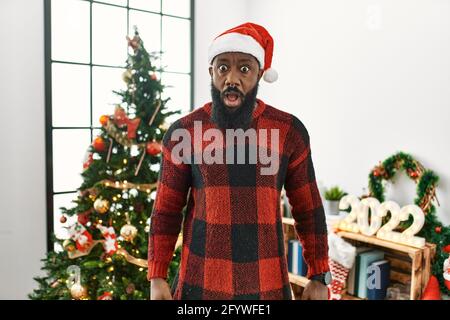 afroamerikanischer Mann mit weihnachtsmannhut, der im Schockgesicht neben dem Weihnachtsbaum steht, skeptisch und sarkastisch aussieht und mit offenem Mund überrascht ist Stockfoto