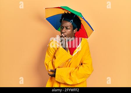 Junger afroamerikanischer Mann mit gelbem Regenmantel gelangweilt gähnend müde, den Mund mit der Hand bedeckend. Unruhig und müde. Stockfoto