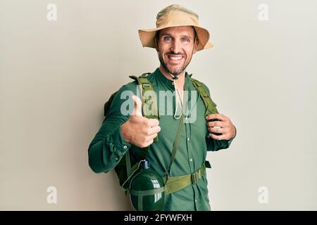 Schöner Mann mit Bart trägt Explorer Hut und Rucksack tun glückliche Daumen nach oben Geste mit der Hand. Genehmigung Ausdruck Blick auf die Kamera zeigt Stockfoto