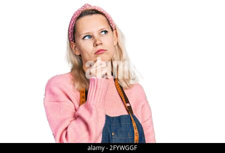 Junge kaukasische Frau Schneiderin Designerin trägt in der Modeatelier Schürze Denken konzentriert Über Zweifel mit Finger auf dem Kinn und Blick nach oben fragen Stockfoto