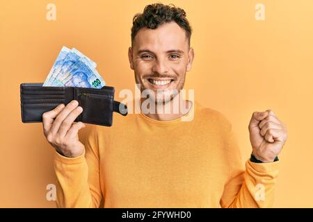 Junger hispanischer Mann hält Geldbörse mit südafrikanischen Rand-Banknoten und schreit stolz, feiert Sieg und Erfolg sehr aufgeregt mit erhobenem Arm Stockfoto