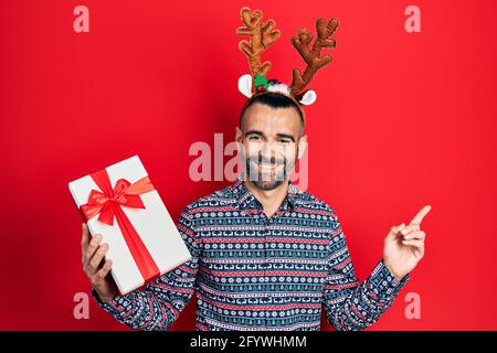 Junger hispanischer Mann trägt Hirsch weihnachtshut mit Geschenk lächelnd Glücklich zeigen mit Hand und Finger zur Seite Stockfoto