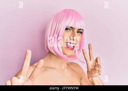 Junger Mann trägt Frau Make-up trägt rosa Perücke lächelnd Blick auf die Kamera zeigt Finger tun Siegeszeichen. Nummer zwei. Stockfoto