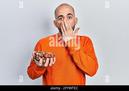 Junger kahler Mann mit rohen Haselnüssen, die den Mund mit der Hand bedecken, schockiert und vor Fehlern fürchtend. Überraschter Ausdruck Stockfoto