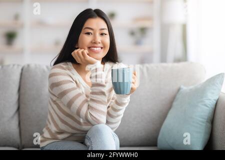 Inländische Freizeit. Glücklich Lächelnde Asiatische Frau Trinkt Kaffee Auf Couch Zu Hause Stockfoto