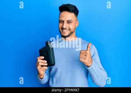 Junger arabischer Mann mit Motorölflasche lächelt glücklich und positiv, Daumen nach oben tun ausgezeichnet und Genehmigung Zeichen Stockfoto