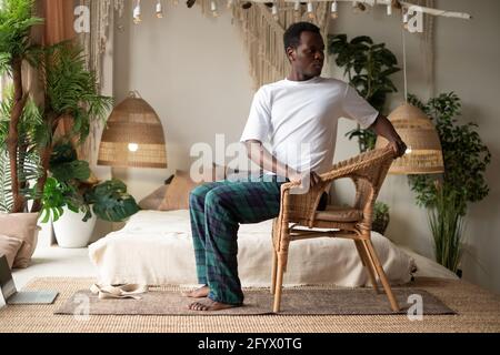 Sportlicher junger afrikanischer Mann, der mit einem Stuhl Yoga praktiziert, Drehstuhl-Pose macht, Parivrtta Utkatasana Stockfoto