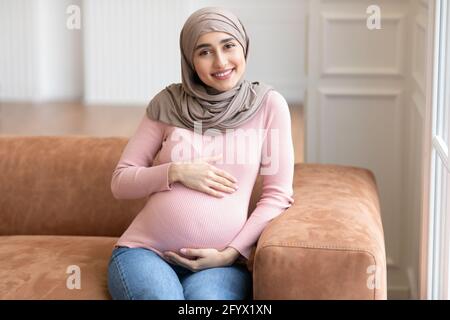 Schwanger Muslim Weiblich In Hijab Sitzt Auf Sofa Zu Hause Stockfoto