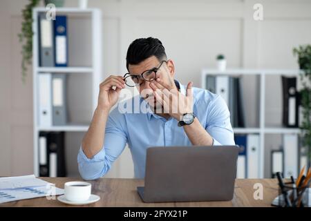 Arabischer Geschäftsmann mit müden, vereiterten Augen, sitzt am Schreibtisch vor dem Laptop, erschöpft von der Online-Arbeit im Büro Stockfoto