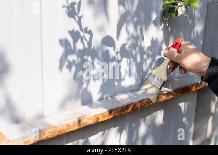 Hand des Mannes hält einen Pinsel und malt über der rostigen Konstruktion mit grauer Farbe. Arbeit im Hinterhof des Hauses. Horizontales Bild. Stockfoto