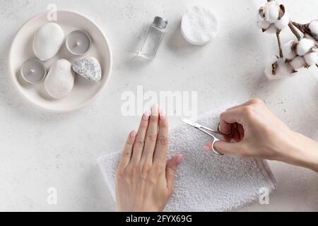 Junge Frau schneidet Nägel mit Maniküre Schere. Hygiene-Konzept. Weißer Hintergrund mit Kerzen und Baumwollblume. Blick von oben. Platz kopieren Stockfoto