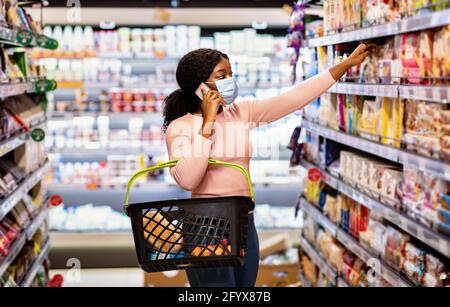 Schwarze Verbraucherin in Maske, die beim Einkauf von Lebensmitteln telefonisch anruft und Produkte aus dem Regal in der Mall nimmt Stockfoto