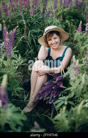 Ein schönes romantisches Mädchen lächelt freudig in einem blauen Kleid und Hut sitzt in einem Feld von lila Lupine Blumen. Weicher, selektiver Fokus. Stockfoto