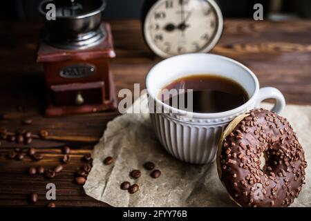 Ein Glas Kaffee auf dem Tisch mit Uhr am Morgen. Ein Vintage-Wecker, der 9:00 Uhr auf dem Tisch neben einer Tasse Kaffee und einem Donut anzeigt. Frühstück. Zeit m Stockfoto