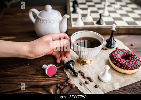 Die Hand hält eine weiße Tasse Kaffee. Schachbrett und verstreutes Schach auf Holztisch. Schokoladendonut für das Frühstück. Brettspiel concep Stockfoto