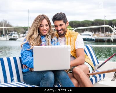 Lächelndes, multiethnisches Paar, das auf Netbook im Internet surft, während es darauf sitzt Motorboot auf Stadtfluss am Tag Stockfoto