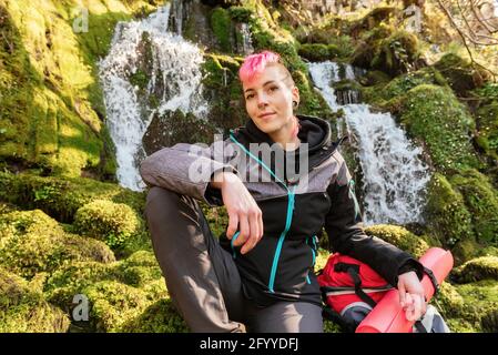 Inhalt weibliche Wanderer mit gefärbten Haaren Blick auf die Kamera während Sitzen auf moosigen Felsen mit Rucksack in der Nähe von Wasserfall fallen aus Felsenklippe Stockfoto