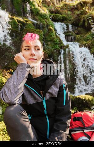 Inhalt weibliche Wandererin mit gefärbten Haaren, die beim Sitzen wegschauen Auf moosigen Felsen mit Rucksack in der Nähe des Wasserfalls, der von felsigen fällt Klippe Stockfoto