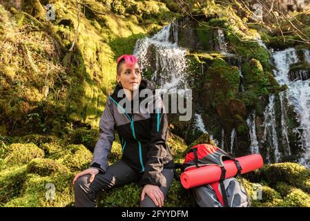 Inhalt weibliche Wandererin mit gefärbten Haaren, die beim Sitzen wegschauen Auf moosigen Felsen mit Rucksack in der Nähe des Wasserfalls, der von felsigen fällt Klippe Stockfoto