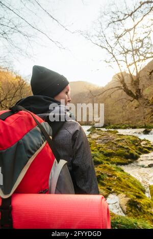 Rückansicht Entdecker mit Rucksack auf moosigen Felsen in der Nähe stehen Schneller Wasserstrom, der durch den Wald mit blattlosen Bäumen fließt Stockfoto