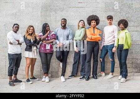 Lächelnde selbstbewusste multirassische Partner in trendigen Bekleidung und Sneakers Stehen auf einem Gehweg in der Stadt auf grauem Hintergrund Stockfoto
