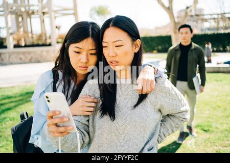 Ernsthafte junge ethnische Freundinnen kuscheln beim Ansehen von Videos Mobiltelefon, das auf dem Rasen im Park in der Nähe eines anonymen Mannes steht Stockfoto