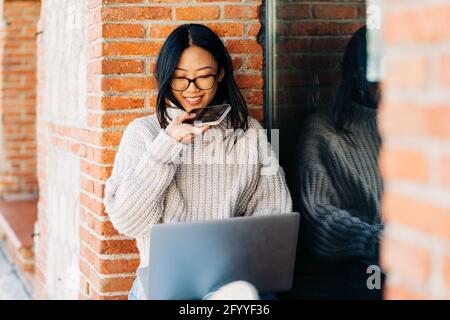 Glückliche junge asiatische Freiberuflerin mit langen dunklen Haaren in Stilvoller Pullover und Brillen, die lächeln und gleichzeitig Sprachnachrichten aufzeichnen Remote arbeiten o Stockfoto