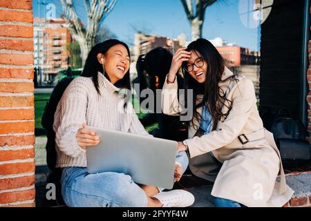 Fröhliche junge asiatische weibliche beste Freunde mit langen dunklen Haaren In stilvollen Outfits lachen, während Sie lustige Videos auf dem Laptop ansehen In der Nähe eines Ziegelbaus Stockfoto