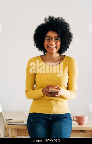 Glückliche afroamerikanische Frau mit Handy lächelnd und aussehend An der Kamera neben dem Tisch mit Tasse und Notizbuch Stockfoto