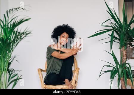 Voller Körper von glücklichen afroamerikanischen Frau lächeln und verwenden Mobiltelefon, während Sie zu Hause Selfie auf einem Holzstuhl machen Stockfoto