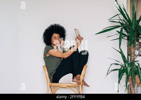 Voller Körper von glücklichen afroamerikanischen Frau lächeln und verwenden Mobiltelefon, während Sie zu Hause Selfie auf einem Holzstuhl machen Stockfoto