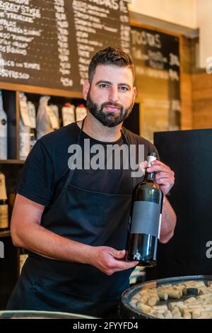 Ernsthafter bärtiger Barkeeper mit dunklem Haar in schwarzer Schürze stehend An der Theke und Vorführung einer Flasche Rotwein während der Arbeit Im Restaurant Stockfoto