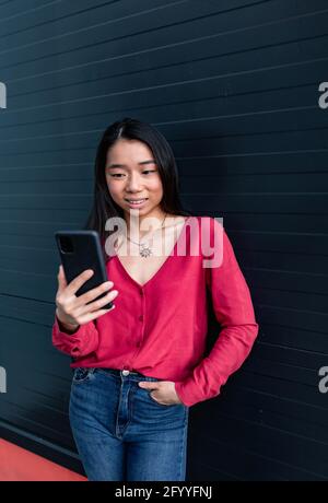 Lächelnde ethnische Frau in legerer Kleidung, die während des Besuchs auf dem Mobiltelefon surft Stehend mit der Hand in der Tasche der Jeans Stockfoto