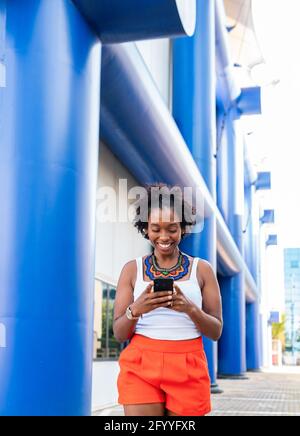 Fröhliche junge afroamerikanische Frau in Sommershorts, die auf dem Handy surfen Telefonieren und auf der sonnigen Straße spazieren gehen Stockfoto
